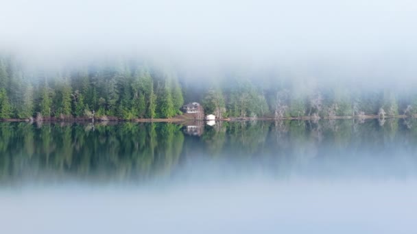 鏡に映る永遠の緑の森の平和的な景色はまだ水面を反映しています 低霧雲の山林の風景のウォーターフロントの木製キャビン シネマティックな空中山岳湖の風景 — ストック動画