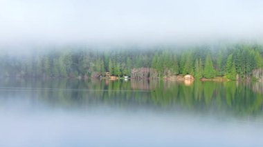 Dağ ormanlarında, alçak sis bulutu içinde rıhtım kenarındaki ahşap kulübeler. Sinema havası dağ gölü manzarası. Durgun su yüzeyine yansıyan ebedi yeşil ormanın huzurlu manzarası