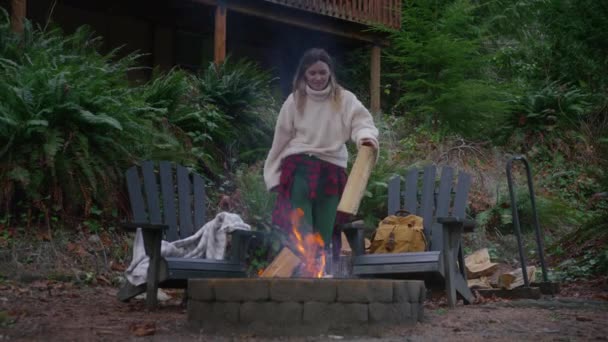 一个年轻的女人在木屋外面的火坑里烧木柴 在绿林4K的熊熊烈火中 女人们在村舍里放松自己 美国偏远自然目的地的周末出游 — 图库视频影像