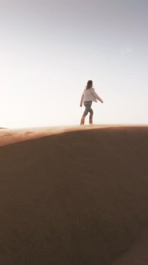 Dikey video. Kum tepesinin zirvesinde duran bir insan aşağıdaki engin çöl manzarasını inceliyor. Rüzgâr saçlarında dalgalanır Geniş bir bakış açısıyla.