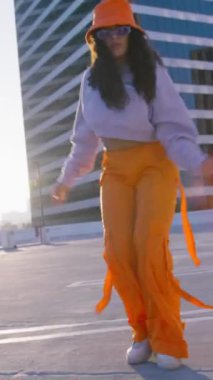 Dikey video. Neşeli ve mutlu kıvırcık saçlı genç kadın altın günbatımı arka planında çatıda dans ediyor. Modaya uygun turuncu pantolon ve kova şapkalı çok ırklı dansçı..