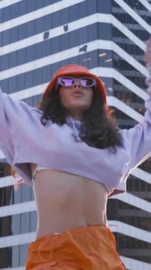 Dikey video. Açık havada kadın dansçı serbest stil hareketlerle dans ediyor. Komik stil sahibi çok ırklı hippi kız. Modayı takip eden güneş gözlüklü ve dışarıda dans eden turuncu şapkalı kadın.