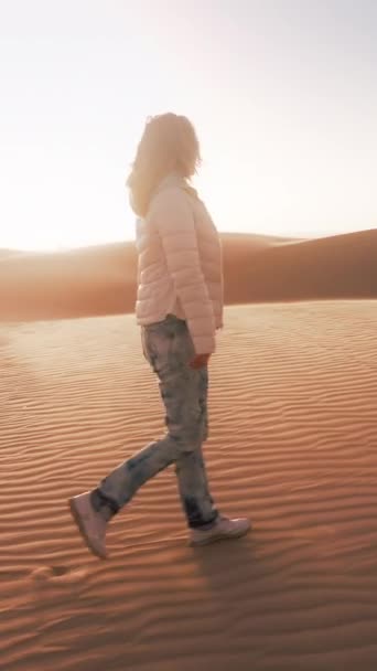 垂直录像 一个女人正在穿越一个被沙子覆盖的沙漠景观 周围都是美丽的沙丘 她正稳步地在广阔干旱的土地上行走 留下了脚印 — 图库视频影像