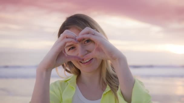 在海滩上美丽的夕阳西下 一位穿着鲜艳的黄色衬衫的快乐的年轻女子手握着一个心形标志 象征着爱情和幸福 4K镜头 图库视频片段