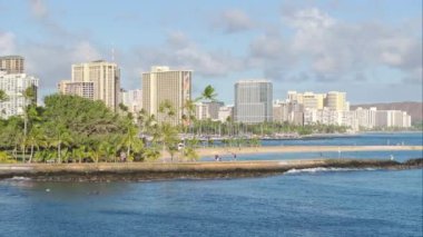 Waikiki 'nin geniş bir görüntüsü, Oahu, Hawaii, ABD' de canlı bir şehir, ışıldayan suyun ötesinden görülüyor. Gökdelenler, palmiye ağaçları ve hareketli sokaklar uzaktan görülebiliyor..