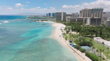 Havadan bakınca Oahu, Hawaii 'deki Waikiki Sahili' nin kumlu kıyı şeridini, turkuaz suları ve çevresindeki binaları görüyoruz. Kamera hareketli sahil bölgesini yüksek katlı kameralarla tarıyor.