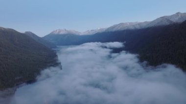 Havadan insansız hava aracı yoğun sis bulutunu yakalıyor tepelerinde dağ zirveleri olan sakin bir gölü örtüyor. Dinginlik, sabahın erken saatlerinde sakinliğe hitap eder. 4K görüntü. 