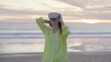 Sanal bir çevreye dalmış, sarı tişörtlü genç bir kadın plajda duruyor, bir VR kulaklık takıyor, çarpıcı bir günbatımının arka planına karşı yeni dünyalar keşfediyor. 4K görüntü. 
