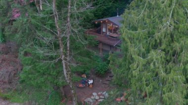 Washington, ABD 'de bir nehrin yakınındaki bir ormanda bulunan ahşap bir kulübeyi araştırın. Ateşin yanında oturan genç bir çifte tanık olun, doğal çevrenin tadını çıkarıyorlar. Havadan. 4K görüntü.