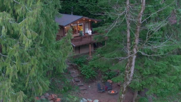 小木屋森林中靠近河流的一个木制小木屋 旁边有一个火坑在燃烧一对年轻夫妇站在阳台上欣赏风景 华盛顿Washington 4K镜头 — 图库视频影像