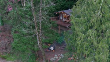 Nehir kenarındaki ağaçlarla çevrili ormandaki ahşap kulübeyi ele geçirin. Genç bir çift ateşin yanında oturur, doğal çevrenin tadını çıkarırlar. - Washington. ABD. Havadan. 4K görüntü.