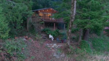 Bir kız, sakin bir ormanın kalbine yerleşmiş, rahat bir ahşap evin dışında kamp ateşinin yanında dinleniyor. Alacakaranlık, nehrin kenarına yerleşiyor. - Washington. ABD. Havadan. 4K görüntü.