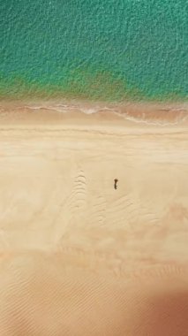 Soluk kesen 4K hava videosundan keyif alan bir adam, turkuaz su ile kumlu sahilde koşuyor ve cep telefonunda dikey görüş için mükemmel. Kıyı sahnesini yüksek çözünürlükte yakala