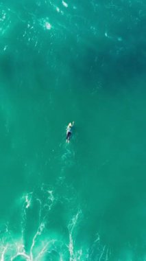 Dikey ekran: berrak turkuaz sularda sörf tahtasında kürek çeken, güzel bir okyanus ortamında huzurlu bir anı yakalayan, huzur ve rahatlama çağrısında bulunan bir insanın sakin bir sahnesi