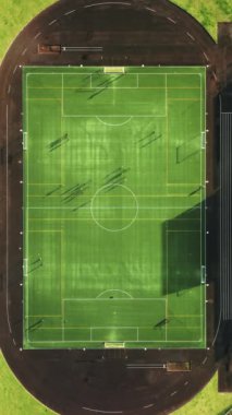 Dikey Ekran: Eğitimdeki oyuncuların olduğu bir futbol sahasını görüntüler. Çimenlikteki güneş ışığı gölgeleri, stadyumdaki dinamik antrenman oturumunu canlandıran canlı bir desen oluşturur.