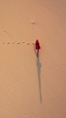 Dikey Ekran: Kırmızı elbiseli bir kız engin çölde tek başına yürürken görülüyor, kumlu arazide güzel bir kontrastlık yaratıyor. Dikey formatta havadan çekilen bir görüntü..