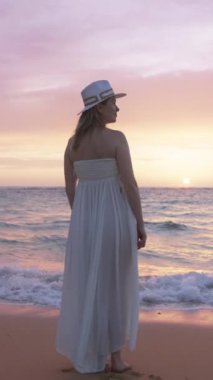 Beyaz elbiseli ve şapkalı bir kadın gün batımında sahilde duruyor, renkli gökyüzüne ve okyanusa hayran. Huzurlu sahne huzur ve rahatlama yayar, tefekkür ve meditasyon için mükemmeldir.