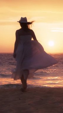 Sahilde gün batımında beyaz elbiseli ve şapkalı romantik bir sahil manzarası seyreden bir kadın hayal edin. Sahil manzarasının dingin güzelliğini korumak için dikey video biçiminde kaydet