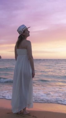 Beyaz elbiseli ve şapkalı bir kadın Hawaii plajında günbatımını seyrediyor, doğada huzurlu bir yaz tatilini somutlaştırıyor. Sahne, okyanus dinginliğini ve sahil güzelliğini yakalıyor.