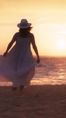 Dikey Ekran: Beyaz elbiseli ve şapkalı bir kadın gün batımında sakin bir dikey videoda sahilde yürüyor. Günbatımı renkleri ve okyanus dalgaları dinlenmek ve eğlenmek için mükemmel bir ortam yaratır.