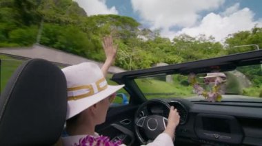 Üstü açık şık bir arabada, Oahu 'da güneşli bir tünelden geçerken neşeyle el sallayan bir kadın. Günlük kıyafetleri yaz günü için mükemmel. 4K görüntü. 