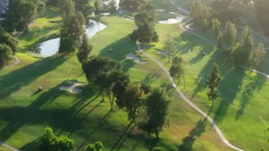 Hava 4K görüntüsü gündoğumunda sakin bir golf sahası yakalar, yemyeşil çayırlar, kum sığınakları ve oyuncularla birlikte huzurlu ve güzel manzarada bir maçın keyfini çıkarırlar.
