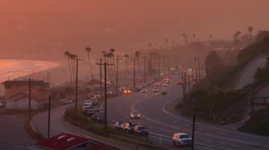 Malibu, California 'nın büyüleyici güzelliğini gün batımında yukarıdan yakalıyor, sahil yolu, palmiye ağaçları ve sakin akşam atmosferini sunuyor, resim gibi ve rahatlatıcı bir manzara sağlıyor.