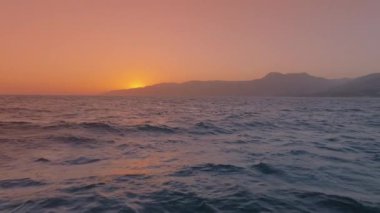 Pelican 'ların yavaşça süzülüşünü izleyin. Okyanusun kenarındaki renkli bir günbatımına karşı. Büyüleyici 4K ile. Sakin dalgalar ve sakin bir atmosfer için uzak dağlarla huzurlu bir ortamın tadını çıkar.