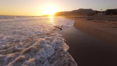 Malibu Sahili, Kaliforniya 'da gün batımını izleyin. Bir FPV insansız hava aracı tarafından ele geçirildi. Rengarenk bir gökyüzü altında sakin bir okyanus ve kıyı görüntüsü sunuyor.