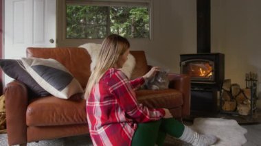 Huzurlu bir aile ortamında, sakin bir sahne sevgili kedisini şöminenin sıcak, davetkar ve rahatlatıcı bir atmosfer yaratan sıcacık ışığıyla okşayan bir kadını gözler önüne serer.