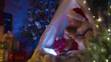 Büyülü bir Noel ortamında, bir anne kızına bayram dekoruyla süslenmiş rahat bir çadırın altında el fenerleriyle kitap yakarak neşeli bir hikaye okur. 4K görüntü. 