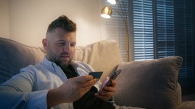 Bir adam dikkatlice kredi kartı bilgilerini güvenli bir online alım için telefonuna girmeye odaklanır. İşlem sürecinin sorunsuz ve sorunsuz geçmesini sağlar.