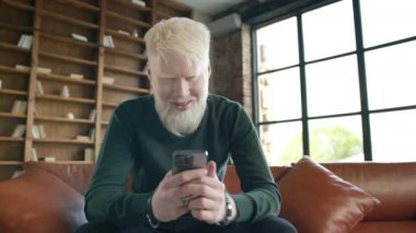 Young smiling albino man holding smartphone device texting sms message sitting at home office. Positive guy using apps playing mobile game chatting in social media surfing web on phone in modern home