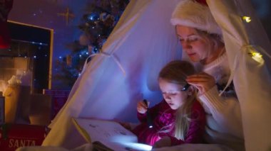 Bir anne, yılbaşı süsleriyle çevrili sıcak bir çadırda genç kızına nazikçe bir Noel hikayesi okur ve kitabı el fenerleriyle aydınlatır. 4K görüntü. 