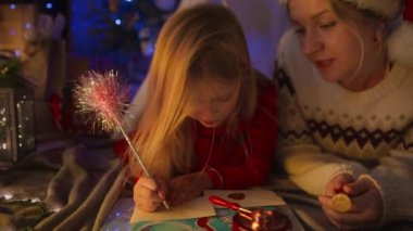 Samimi bir şenlik ortamında, kırmızı elbiseli genç bir kız Noel Baba 'ya mektup yazıyor, annesinin kışın giydiği süveteri giymesine yardım ediyor. Her ikisi de Noel ışıkları ve dekoruyla çevrili.. 