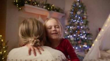 İç açıcı bir Noel sahnesi: bir anne ve kız kucaklaşması, şenlikli süslemeler ve parıldayan bir ağaçla çevrili. 4K görüntü. 