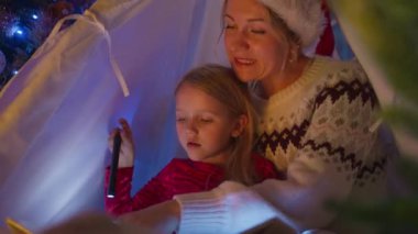 Bir anne ve kızının Noel süslemeleriyle çevrili rahat bir çadırda festival kitabı okumak için el fenerlerini kullandıkları yakın plan bir fotoğraf. 4K görüntü. 