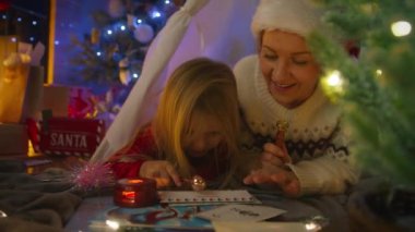 Sıcak bir Noel çadırında, bir anne Noel Baba 'ya Noel süslemeleriyle çevrili bir bayram mektubunu dikkatlice damgalamasında kızına yardım eder. 4K görüntü. 