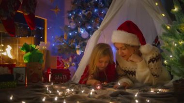 Sıcak bir çadırda yakalanan bir anne, kızının Noel Baba 'ya yazdığı mektubu Noel ışıklarının altında damgalamasına yardım eder. 4K görüntü. 