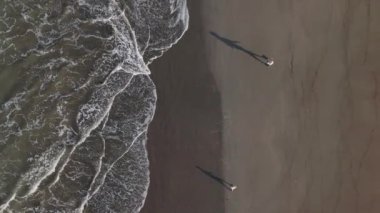 Yukarıdan, bir baba ve oğul kumsalda el ele yürür, siluetleri ılık kumda, yumuşak dalgalar kıyıda süzülürken, barışçıl bir atmosfer yaratarak bağlarını ve neşelerini paylaşırlar.