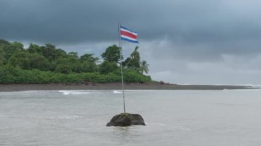 Kosta Rika 'nın çarpıcı ve çarpıcı bir görüntüsü. Canlı bir bayrak, yemyeşil ve sakin sularla çevrili büyük bir kayanın üzerinde gururla sallanıyor.