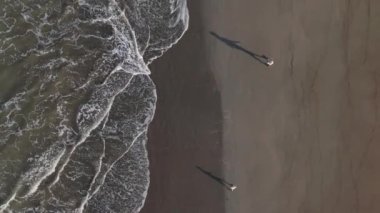 Yukarıdan, bir baba ve oğul kumsalda el ele yürür, siluetleri ılık kumda, yumuşak dalgalar kıyıda süzülürken, barışçıl bir atmosfer yaratarak bağlarını ve neşelerini paylaşırlar.