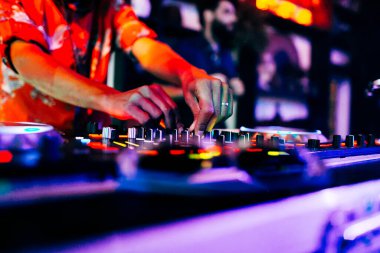 DJ bir gece kulübünde müzik çalıyor. Ellerinin müziği DJ mikseriyle karıştırdığını ve arka plandaki bir partinin rengini görebilirsiniz..