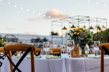 Şarap kadehleri ve çiçekli yemek masası, düğün için mükemmel, zarif bir parti ya da açık hava kutlaması için. Sıcak ışıklar ve yumuşak bokeh samimi ve lüks bir atmosfer yaratır..