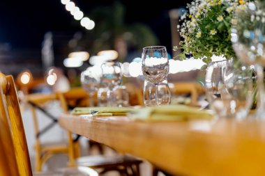 Şarap kadehleri ve çiçekli yemek masası, düğün için mükemmel, zarif bir parti ya da açık hava kutlaması için. Sıcak ışıklar ve yumuşak bokeh samimi ve lüks bir atmosfer yaratır..