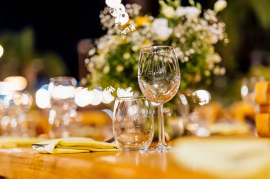 Şarap kadehleri ve çiçekli yemek masası, düğün için mükemmel, zarif bir parti ya da açık hava kutlaması için. Sıcak ışıklar ve yumuşak bokeh samimi ve lüks bir atmosfer yaratır..