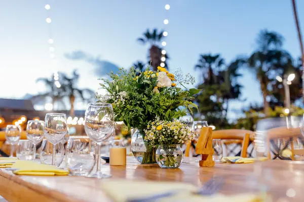 Şarap kadehleri ve çiçekli yemek masası, düğün için mükemmel, zarif bir parti ya da açık hava kutlaması için. Sıcak ışıklar ve yumuşak bokeh samimi ve lüks bir atmosfer yaratır..