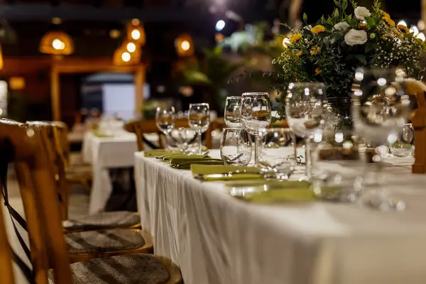 Şarap kadehleri ve çiçekli yemek masası, düğün için mükemmel, zarif bir parti ya da açık hava kutlaması için. Sıcak ışıklar ve yumuşak bokeh samimi ve lüks bir atmosfer yaratır..