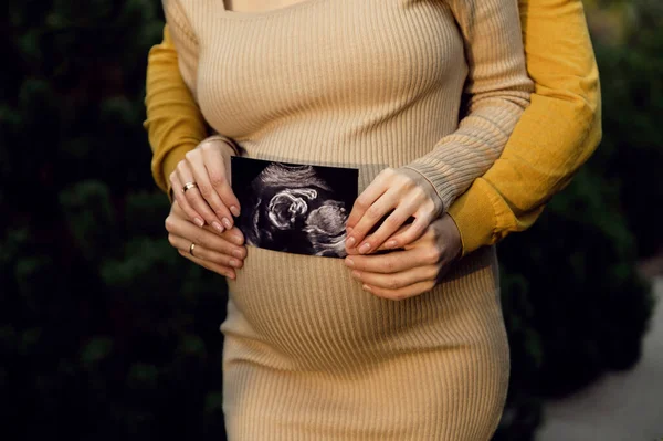 a pregnant couple holds an ultrasound picture on the background of the abdomen. close-up of a pregnant belly. 4d ultrasound of the fetus.modern ultrasound picture.