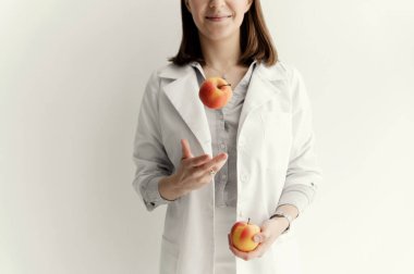 Beyaz arka planda beyaz önlüklü, gülümseyen doktor beslenme uzmanı eline elma fırlatıyor.